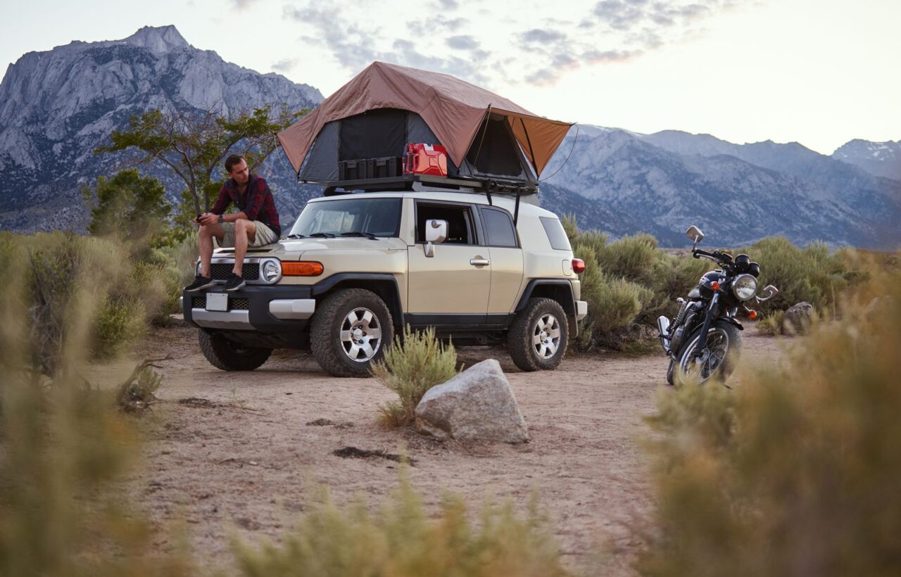 Beige SUV with an open rooftop tent in a desert valley below mountains, man sitting on the hood