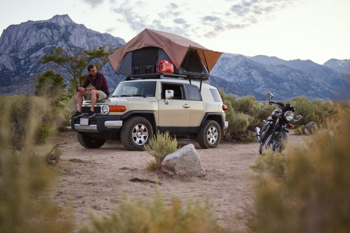 Beige SUV with an open rooftop tent in a desert valley below mountains, man sitting on the hood