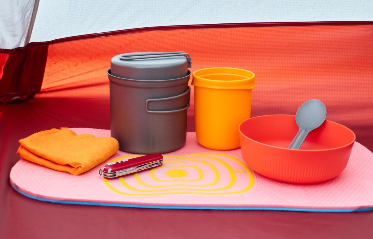 Camp cookware, cup, bowl, towel and pocketknife laid out neatly on a mat inside a red tent