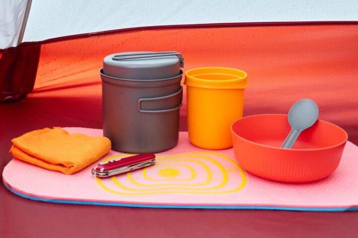 Camp cookware, cup, bowl, towel and pocketknife laid out neatly on a mat inside a red tent