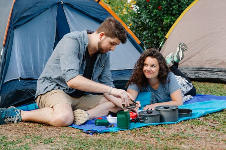 Couple on a picnic blanket in front of dome tents, lighting a camp stove next to cooking pots