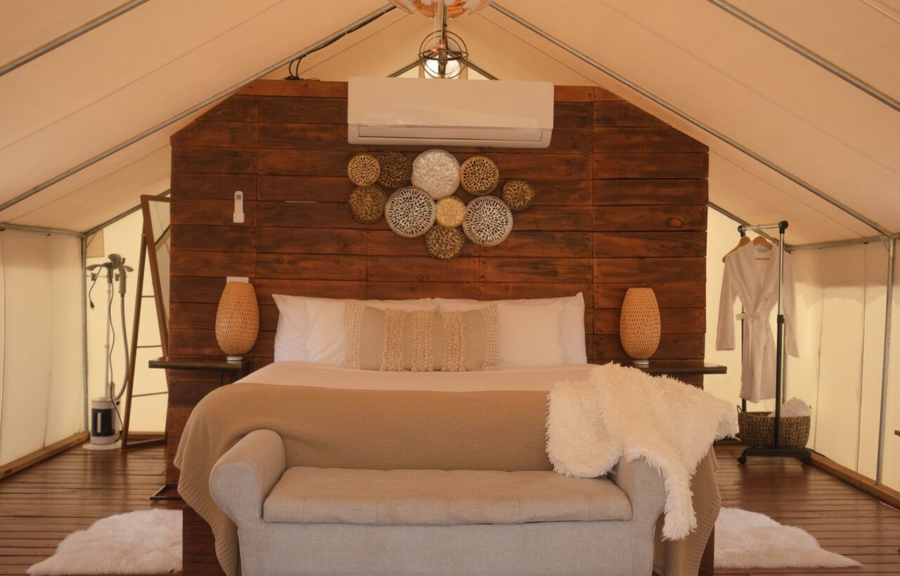Safari-style glamping tent interior with a made bed, wood headboard wall, bench and robe rack