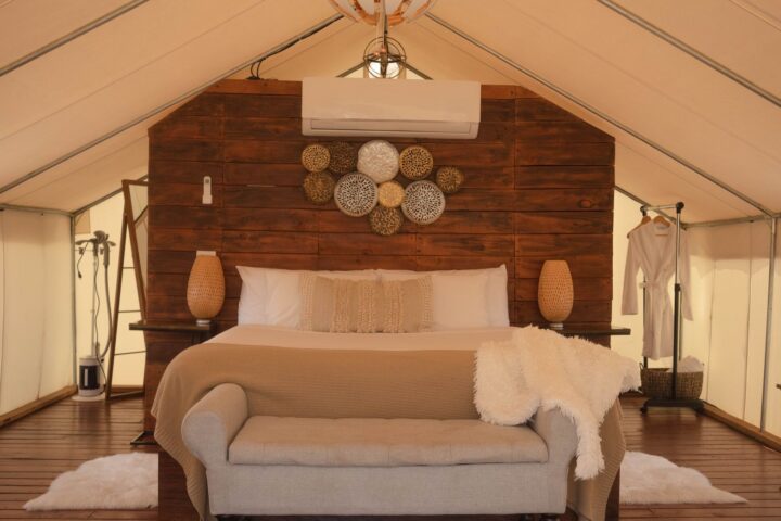 Safari-style glamping tent interior with a made bed, wood headboard wall, bench and robe rack