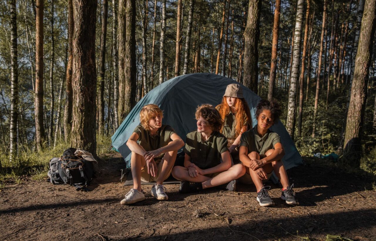 Four children in matching outdoor shirts sitting in front of a blue dome tent in a pine forest