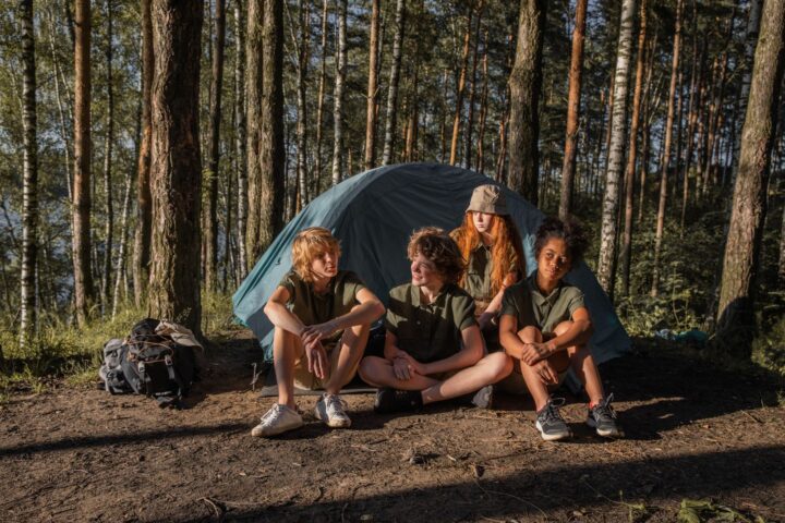 Four children in matching outdoor shirts sitting in front of a blue dome tent in a pine forest
