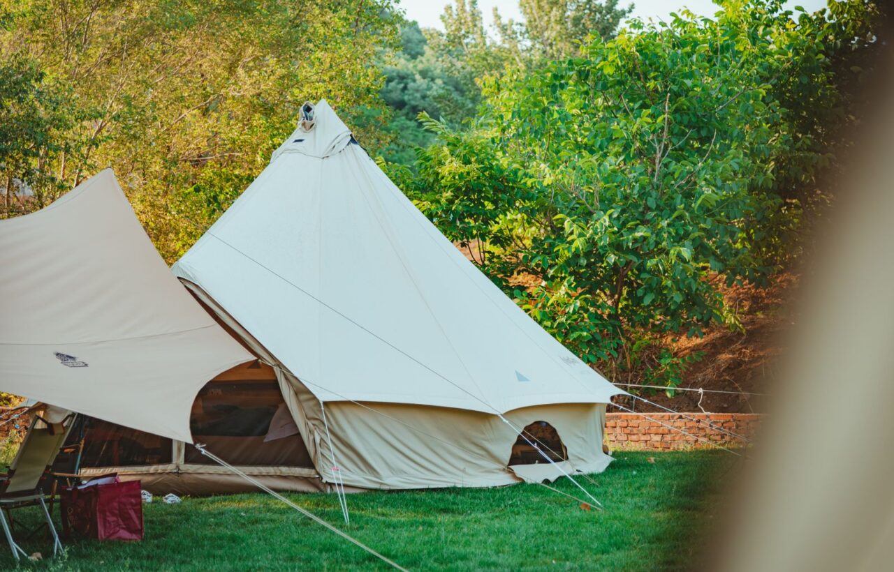 Cream canvas bell tent pitched on a green lawn with guy lines, next to a tarp shelter and trees