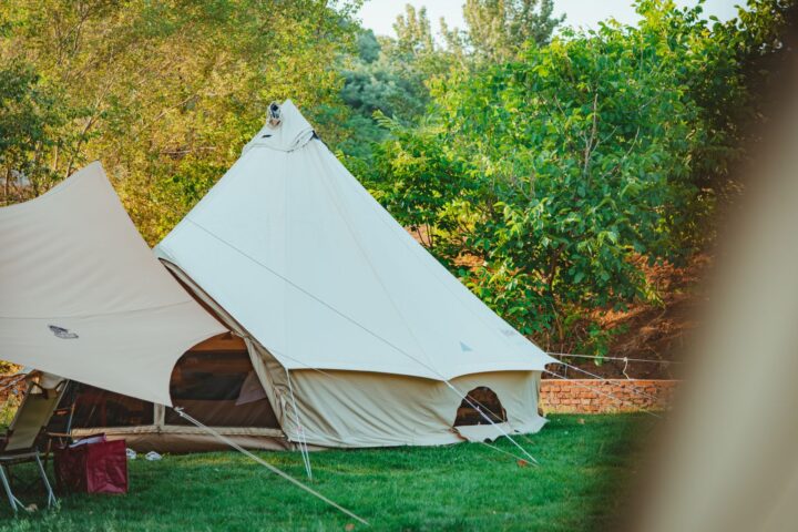 Cream canvas bell tent pitched on a green lawn with guy lines, next to a tarp shelter and trees