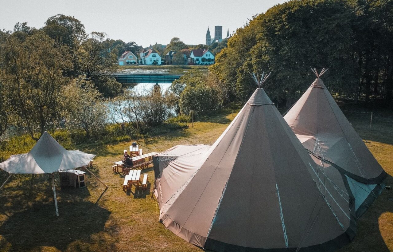Two large canvas tipi tents and picnic tables on a riverside lawn, town and church spire behind