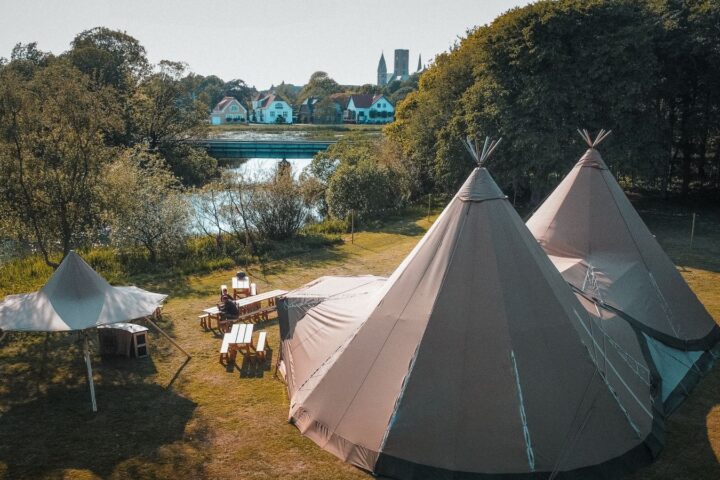 Two large canvas tipi tents and picnic tables on a riverside lawn, town and church spire behind