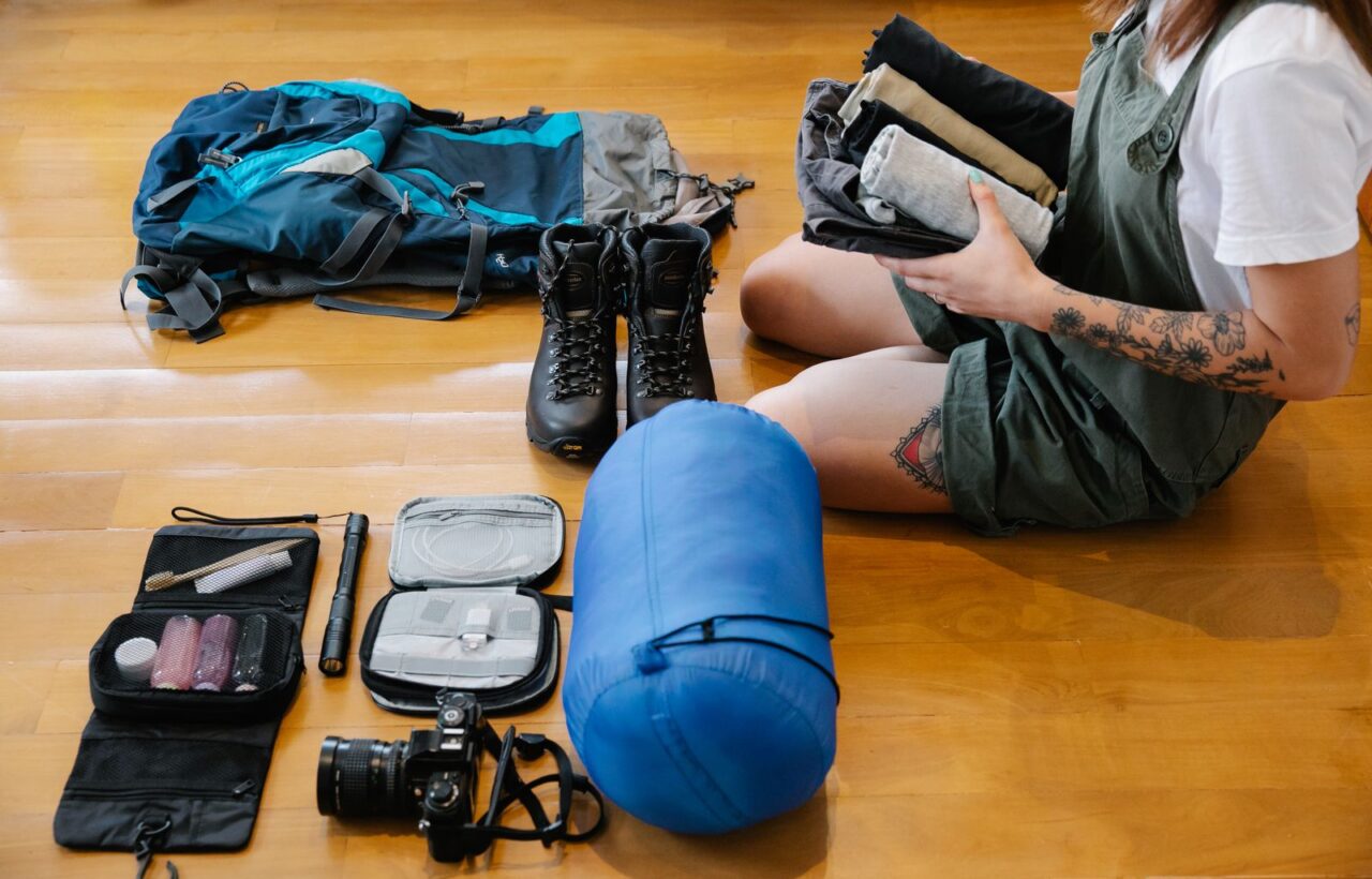 Person holding rolled clothes beside a backpack, hiking boots, sleeping bag and toiletry kit on a floor