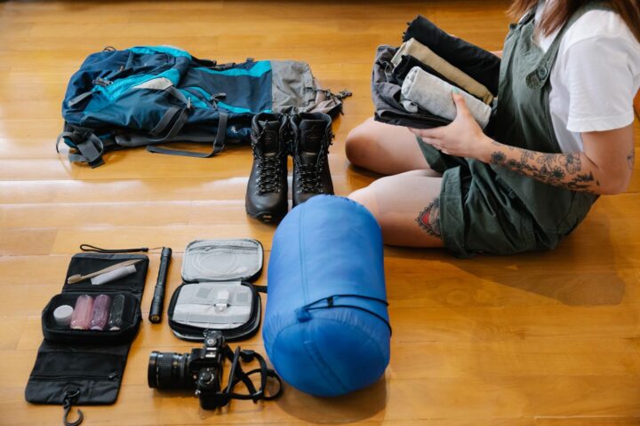 Person holding rolled clothes beside a backpack, hiking boots, sleeping bag and toiletry kit on a floor
